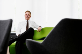 Sitzendes Mitarbeiterportrait eines Ausbilders in moderner Besprechungsumgebung mit zwei Lounge Stühlen im unscharfen Vordergrund