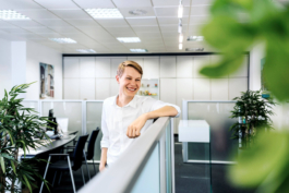 Stehendes Mitarbeiterportrait eines Auszubildenden in modernem Großraumbüro. Auszubildender lehnt lässig an einer Trennwand