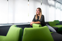 Sitzendes Mitarbeiterportrait einer Auszubildenden in modernem Wartebereich eines Unternehmens. Auszubildende sitzt lächelnd vor Fensterfront.