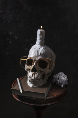 Produktfoto. Ein Skeletierter Schädel mit brennender Kerze trägt eine hochwertige, goldene Brille. Um ihn herum liegen Zeichenbuch, Stift und zerknülltes Papier.