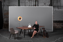 Produktfotografie eines Tisches. Eine reich wirkende Dame sitzt zusammen mit ihrem Jagdhund an ihrem Esstisch und schaut teilnahmslos. Im Hintergrund steht eine deplaziert wirkende graue Wand.