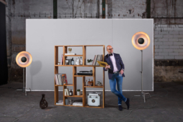 Produktfotografie eines Holzregals. Ein geschmackvoll gekleideter Mann lehnt an das Regal welches mit Genussmitteln und Utensilien seiner Hobbys gefüllt ist. Im Hintergrund steht eine deplaziert wirkende hellgraue Wand.