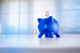 Stilllifefotografie eines blauen Sparschweins vor hochwertigem Hintergrund in das eine Geldmünze fällt.