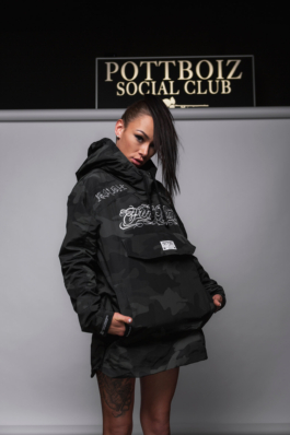 Werbefotografie einer Tarnfarbenen Jacke. Eine tätowierte Frau steht vor grauem Hintergrund. Über dem Hintergrund sieht man das Firmenlogo als Leuchtkasten.