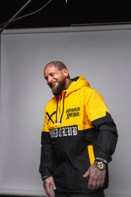 Werbefotografie einer Tarnfarbenen Jacke. Ein stark tätowierter Mann steht vor grauem Hintergrund dessen Enden man klar im Bild erkennen kann. Im oberen Bereich kann man Teile eines Lampenstativs und ein Kabel erkennen..