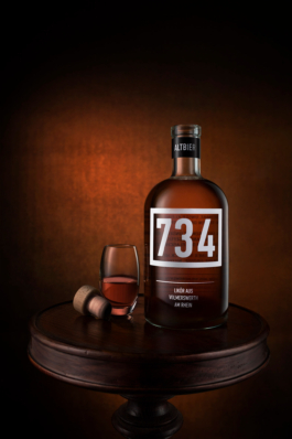 Werbefotografie einer Spirituosenflasche der Marke 734 vor edlem undefiniertem rötlichen Hintergrund. Neben der Flasche steht ein befülltes Likörglas und ein Korken liegt seitlich daneben.