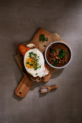 Foodfotografie eines Schnitzels mit Ei auf einem Holzbrettchen vor Betonhintergrund. Neben dem Brettchen liegt eine kleine Schaufel mit Meerzalz.