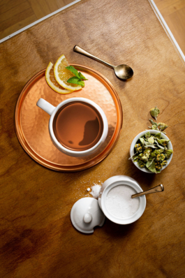 Produktfotografie einer Teesituation auf einem Coffeetable. Neben der gefüllten Teetasse sind Teeblatter, Zucker und Zitronenscheiben angeordnet.