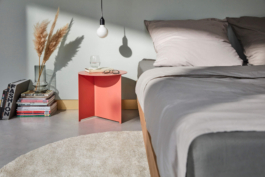 Produktfotografie eines aprikot farbenen Beistelltisches in modernem Schlafzimmer. Auf dem Beistelltisch der Marke Feli steht stylische Deko. Sonneneinfall durch ein Fenster erzeugt eine stimmungsvolle Dramatik.