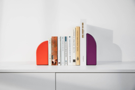 Produktfotografie von Buchstützen der Marke FELI in moderner Location. Geometrische Buchstützen in den Farben Dunkelmagenta und Aprikot stützen eine Reihe Bücher.